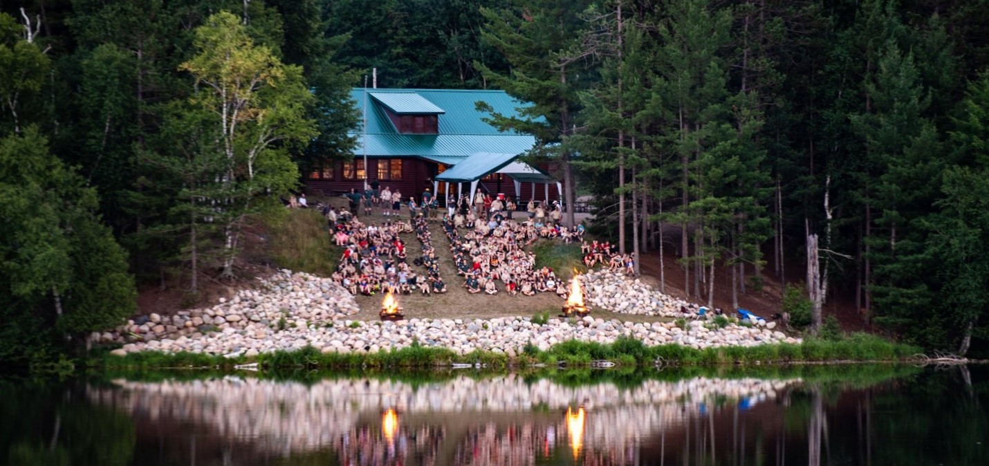 Scouts gather on the shores of Lake Killian, watching two large camp fires roar in front of the lake, with the Dining Hall in view on the hill above them.